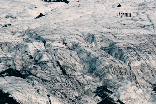 A vast, rugged glacier landscape with noticeable crevasses, showing a group of people standing in the distance on the glacier surface. The ice has a textured, layered appearance with dark ridges running through it.