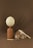 A still life composition featuring a large egg or round object on top of a slender, earthy-toned vase. Next to it, there is a tall, patterned feather leaning against a rustic loaf of bread. In front lies a vintage fork and a butter knife with cream-colored handles.