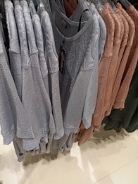 A clothing rack displaying a collection of knitted sweaters in varying colors including light gray, brown, and green. The sweaters are arranged neatly on black hangers, with a visible clothing tag hanging from one of them, indicating a brand or size. The setting appears to be a retail store with a clean, tiled floor.