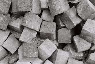 A collection of numerous grey cobblestones piled together, creating a textured and rough surface. The stones are irregularly shaped but generally follow a square-like form, showcasing a rugged and industrial aesthetic.