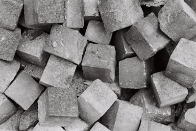 A collection of numerous grey cobblestones piled together, creating a textured and rough surface. The stones are irregularly shaped but generally follow a square-like form, showcasing a rugged and industrial aesthetic.