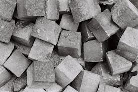 A collection of numerous grey cobblestones piled together, creating a textured and rough surface. The stones are irregularly shaped but generally follow a square-like form, showcasing a rugged and industrial aesthetic.