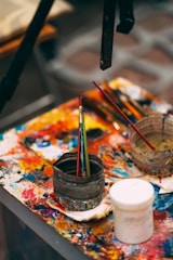 A cluttered artist's workspace featuring a table covered with colorful, smeared paint. Various paintbrushes are placed inside a rugged container, alongside other painting tools. The vividly splattered colors provide a sense of creative chaos.
