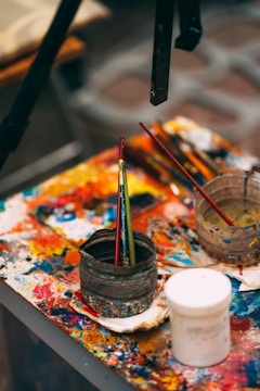 A cluttered artist's workspace featuring a table covered with colorful, smeared paint. Various paintbrushes are placed inside a rugged container, alongside other painting tools. The vividly splattered colors provide a sense of creative chaos.