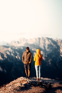 Two individuals dressed in outdoor clothing stand on a rocky outcrop with a mountainous landscape in the background. One wears a yellow jacket, and the other a dark jacket and shorts. A backpack is lying on the ground nearby, suggesting a hiking trip.