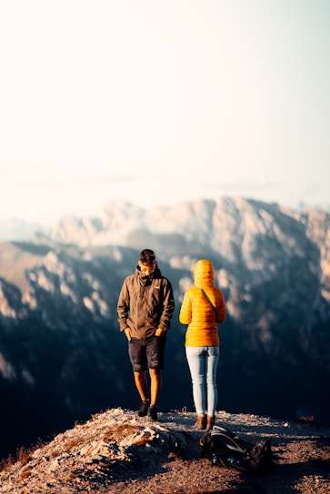 Two individuals dressed in outdoor clothing stand on a rocky outcrop with a mountainous landscape in the background. One wears a yellow jacket, and the other a dark jacket and shorts. A backpack is lying on the ground nearby, suggesting a hiking trip.