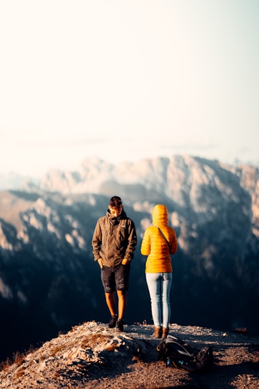 Two individuals dressed in outdoor clothing stand on a rocky outcrop with a mountainous landscape in the background. One wears a yellow jacket, and the other a dark jacket and shorts. A backpack is lying on the ground nearby, suggesting a hiking trip.