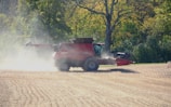 A red combine harvester is working in a field, surrounded by trees with dense green foliage. Dust is billowing around the machine as it moves across the recently harvested ground, indicating a dry and sunny day. The background shows a forested area under bright sunlight.