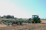 A large green tractor stands on a freshly plowed field, equipped with an industrial disc harrow attachment. The earth is turned, suggesting preparation for planting or plowing. The sky is clear and the surrounding landscape includes distant trees and a few utility poles.