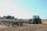 A large green tractor stands on a freshly plowed field, equipped with an industrial disc harrow attachment. The earth is turned, suggesting preparation for planting or plowing. The sky is clear and the surrounding landscape includes distant trees and a few utility poles.