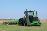 A large green tractor is parked on a grassy field, equipped with farming machinery attached at the back. In the background, there is a vast open field with a distant water tower visible on the horizon.