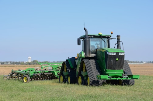 A large green tractor is parked on a grassy field, equipped with farming machinery attached at the back. In the background, there is a vast open field with a distant water tower visible on the horizon.