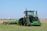 A large green tractor is parked on a grassy field, equipped with farming machinery attached at the back. In the background, there is a vast open field with a distant water tower visible on the horizon.