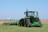 A large green tractor is parked on a grassy field, equipped with farming machinery attached at the back. In the background, there is a vast open field with a distant water tower visible on the horizon.