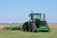 A large green tractor is parked on a grassy field, equipped with farming machinery attached at the back. In the background, there is a vast open field with a distant water tower visible on the horizon.