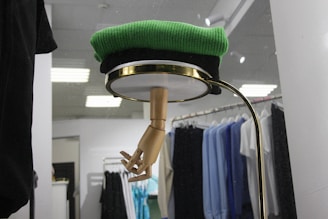A retail display featuring a wooden mannequin hand emerging from the bottom of a circular shelf or glass covering, topped with a folded green sweater. Behind the display, there is a clothing rack with various shirts and garments in black, blue, and white hues, lit by overhead lights.