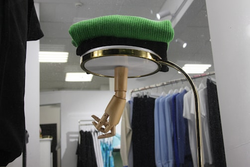A retail display featuring a wooden mannequin hand emerging from the bottom of a circular shelf or glass covering, topped with a folded green sweater. Behind the display, there is a clothing rack with various shirts and garments in black, blue, and white hues, lit by overhead lights.