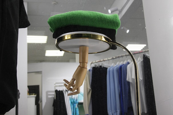 A retail display featuring a wooden mannequin hand emerging from the bottom of a circular shelf or glass covering, topped with a folded green sweater. Behind the display, there is a clothing rack with various shirts and garments in black, blue, and white hues, lit by overhead lights.