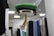 A retail display featuring a wooden mannequin hand emerging from the bottom of a circular shelf or glass covering, topped with a folded green sweater. Behind the display, there is a clothing rack with various shirts and garments in black, blue, and white hues, lit by overhead lights.