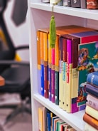 A bookshelf with neatly arranged colorful books. The spines of the books feature a variety of bright colors, including orange, purple, blue, and red. A green tassel hangs over one of the books. The background shows part of an office chair and a desk.