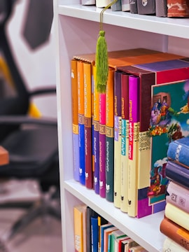 A bookshelf with neatly arranged colorful books. The spines of the books feature a variety of bright colors, including orange, purple, blue, and red. A green tassel hangs over one of the books. The background shows part of an office chair and a desk.