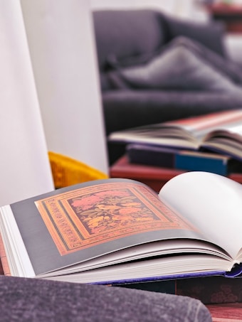 An open art book displaying an intricate floral design with vibrant colors on a page. The book lies on a soft surface in a cozy living room setting with cushions and another blurred book in the background. The color tones in the room are warm and inviting.