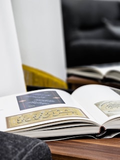 An open book lies on a wooden surface, displaying pages with elegant calligraphy and intricate designs. The background features a blurred sofa and a mirror reflecting another book, creating a cozy and reflective atmosphere.