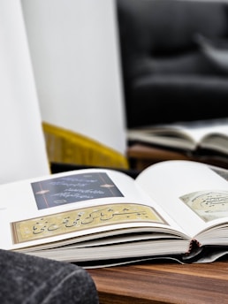 An open book lies on a wooden surface, displaying pages with elegant calligraphy and intricate designs. The background features a blurred sofa and a mirror reflecting another book, creating a cozy and reflective atmosphere.