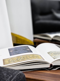 An open book lies on a wooden surface, displaying pages with elegant calligraphy and intricate designs. The background features a blurred sofa and a mirror reflecting another book, creating a cozy and reflective atmosphere.