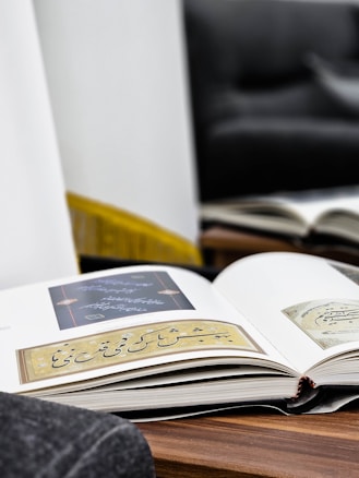An open book lies on a wooden surface, displaying pages with elegant calligraphy and intricate designs. The background features a blurred sofa and a mirror reflecting another book, creating a cozy and reflective atmosphere.