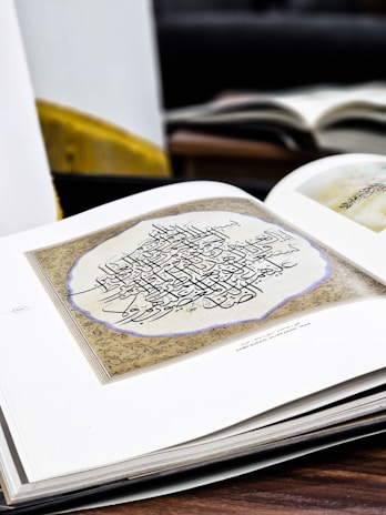 An open book displays an intricately designed piece of calligraphy featuring ornate, black script on a cream page. The text is surrounded by an intricate border with a floral pattern. Other books are visible in the blurred background, hinting at a studious or scholarly setting.