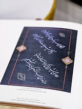 A decorative piece of Persian calligraphy featuring intricate handwriting on a dark background. The borders are accented with red lines and elaborately designed corners incorporating colorful motifs, likely ornamental or floral in nature. Accompanying text at the bottom identifies the work as a poem by Hafiz, composed and arranged in Iran.