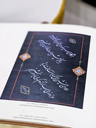 A decorative piece of Persian calligraphy featuring intricate handwriting on a dark background. The borders are accented with red lines and elaborately designed corners incorporating colorful motifs, likely ornamental or floral in nature. Accompanying text at the bottom identifies the work as a poem by Hafiz, composed and arranged in Iran.