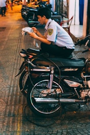 A person wearing a security guard uniform is sitting cross-legged on a motorcycle, absorbed in reading a newspaper. The setting is an urban street at night, with warm lights reflecting off the pavement. Several other motorcycles are parked on the sidewalk in the background.