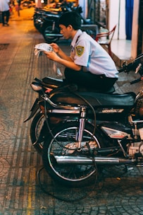 A person wearing a security guard uniform is sitting cross-legged on a motorcycle, absorbed in reading a newspaper. The setting is an urban street at night, with warm lights reflecting off the pavement. Several other motorcycles are parked on the sidewalk in the background.
