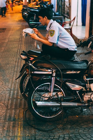 A person wearing a security guard uniform is sitting cross-legged on a motorcycle, absorbed in reading a newspaper. The setting is an urban street at night, with warm lights reflecting off the pavement. Several other motorcycles are parked on the sidewalk in the background.