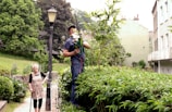 A man is trimming tall plants with an electric hedge trimmer while standing on a platform in a garden. A woman with white hair and a walking stick is standing nearby, smiling. The scene is set in a lush garden with a vintage street lamp, surrounded by greenery, and adjacent to residential buildings.