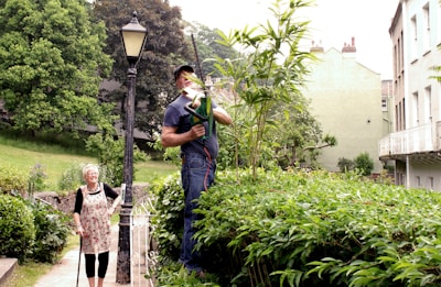 A man is trimming tall plants with an electric hedge trimmer while standing on a platform in a garden. A woman with white hair and a walking stick is standing nearby, smiling. The scene is set in a lush garden with a vintage street lamp, surrounded by greenery, and adjacent to residential buildings.