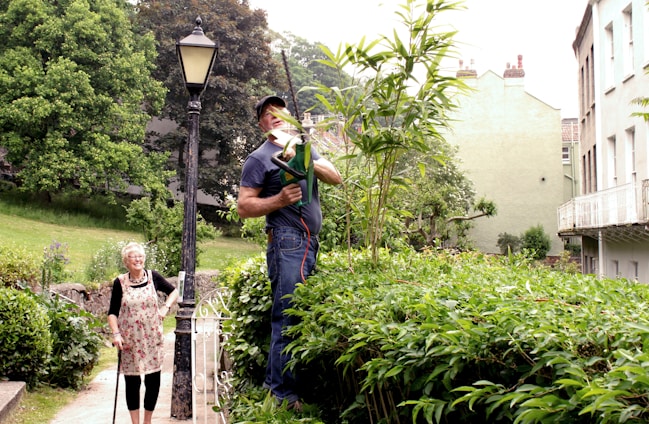 A man is trimming tall plants with an electric hedge trimmer while standing on a platform in a garden. A woman with white hair and a walking stick is standing nearby, smiling. The scene is set in a lush garden with a vintage street lamp, surrounded by greenery, and adjacent to residential buildings.