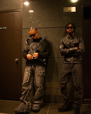Two individuals stand in a dimly lit hallway, dressed in edgy attire. One person wears a puffy jacket over a sweater with baggy, distressed jeans and unique shoes. The other wears a leather jacket and dark sunglasses. The wall behind them is a textured dark color, and the door next to them has a sign labeled 'ELECTRIC ROOM.'