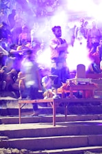 A group of people appears to be gathered outdoors, with a man in traditional attire standing prominently in the center. He is surrounded by smoke, possibly from incense or fire. The background is filled with a crowd of seated and standing individuals, some of whom are blurred, suggesting movement or significant depth. The ambient lighting and haze create a mystical or spiritual atmosphere.