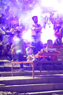 A group of people appears to be gathered outdoors, with a man in traditional attire standing prominently in the center. He is surrounded by smoke, possibly from incense or fire. The background is filled with a crowd of seated and standing individuals, some of whom are blurred, suggesting movement or significant depth. The ambient lighting and haze create a mystical or spiritual atmosphere.