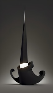 A sculptural, abstract lamp design with a sharp, pointed structure emanating from a coiled, minimalistic base. The primary material appears dark and glossy, with a subtle glow at the center creating a dramatic effect against the muted background.
