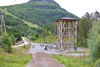 A grassy, forested landscape with a large wooden climbing structure in the foreground. Several people are near the structure and on the path, which winds through the green hills. The scene includes tall trees, power lines, and distant mountain peaks, with a sense of outdoor activity and adventure.