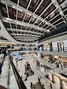 A spacious and modern shopping mall interior with multiple floors. The architecture features a high ceiling with large glass panels. The space is illuminated with bright lights and a large, intricate sculpture hanging prominently.