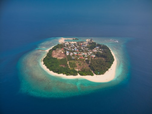A small, lush island surrounded by vibrant turquoise waters and a coastline of white sandy beaches. The island features a dense cluster of buildings and vegetation in the center, with clear water extending into the deep blue ocean.