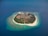 A small, lush island surrounded by vibrant turquoise waters and a coastline of white sandy beaches. The island features a dense cluster of buildings and vegetation in the center, with clear water extending into the deep blue ocean.
