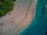 An aerial view of a sandy beach with turquoise waves gently lapping at the shore. A group of people can be seen forming a circle on the sand, surrounded by various beach items such as umbrellas and lounges. The beach is bordered by lush greenery, providing a contrast to the pale sand and vibrant water.
