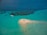 A small, sandy island surrounded by turquoise waters with a group of people standing on it. A white boat is anchored nearby, and darker patches in the water indicate coral or underwater vegetation.