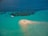 A small, sandy island surrounded by turquoise waters with a group of people standing on it. A white boat is anchored nearby, and darker patches in the water indicate coral or underwater vegetation.